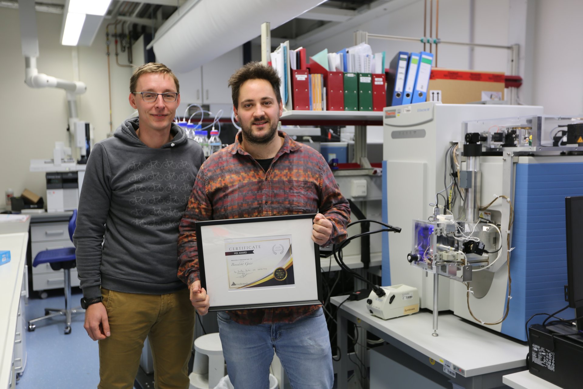 Caption: Awardee Benedikt Geier (right) and Manuel Liebeke in front of the MALDI-MS, which is at the centre of the now award-winning method. (MPIMM/F. Aspetsberger)
